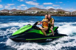 Happy couple wearing life jackets riding a green jet ski across the crystal-clear waters of Lanzarote, with volcanic mountains and the island's coastline in the background.