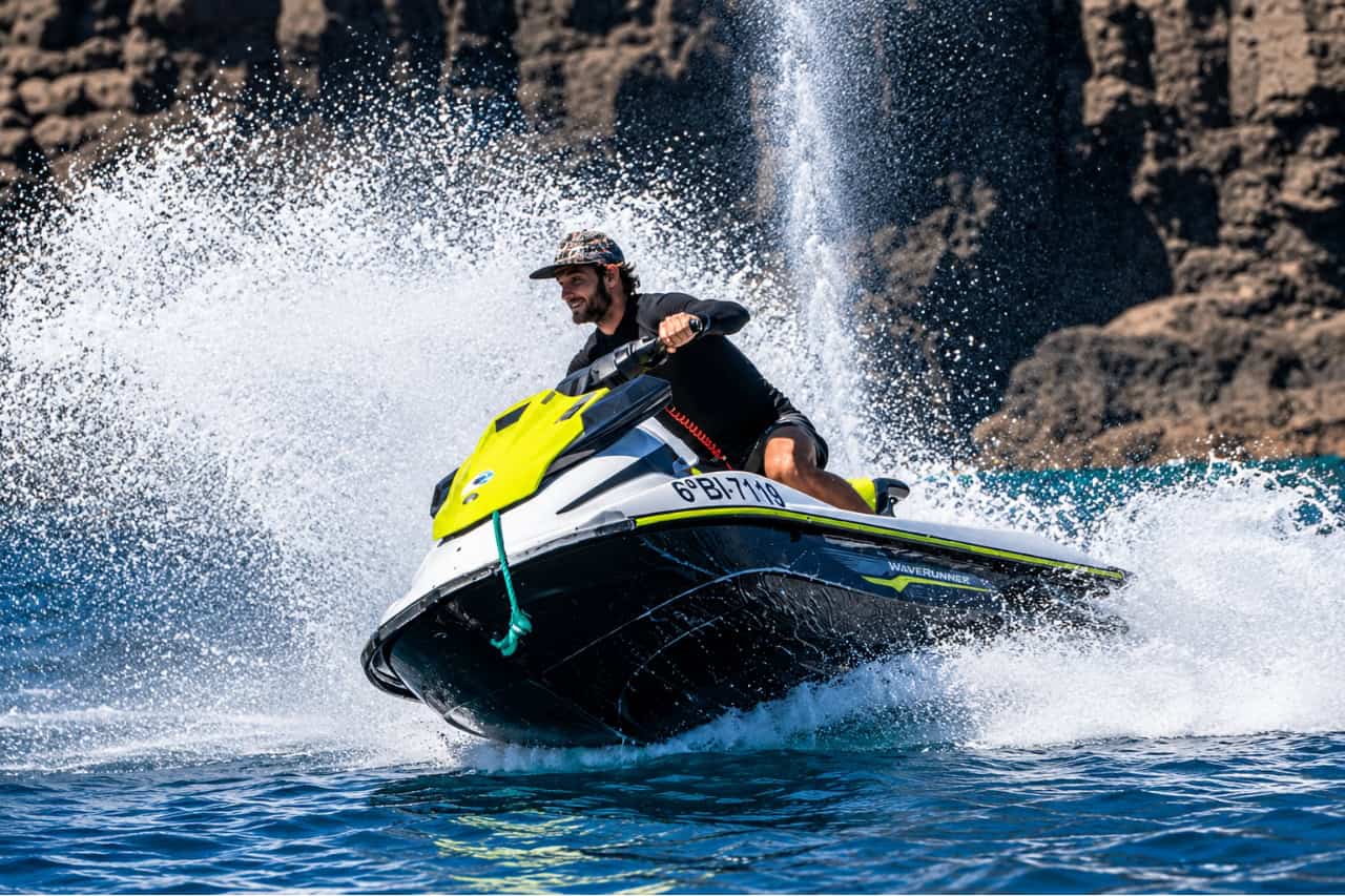 Person riding a jet ski at high speed off the coast of Puerto del Carmen, Lanzarote, creating a large spray with dramatic volcanic cliffs in the background.