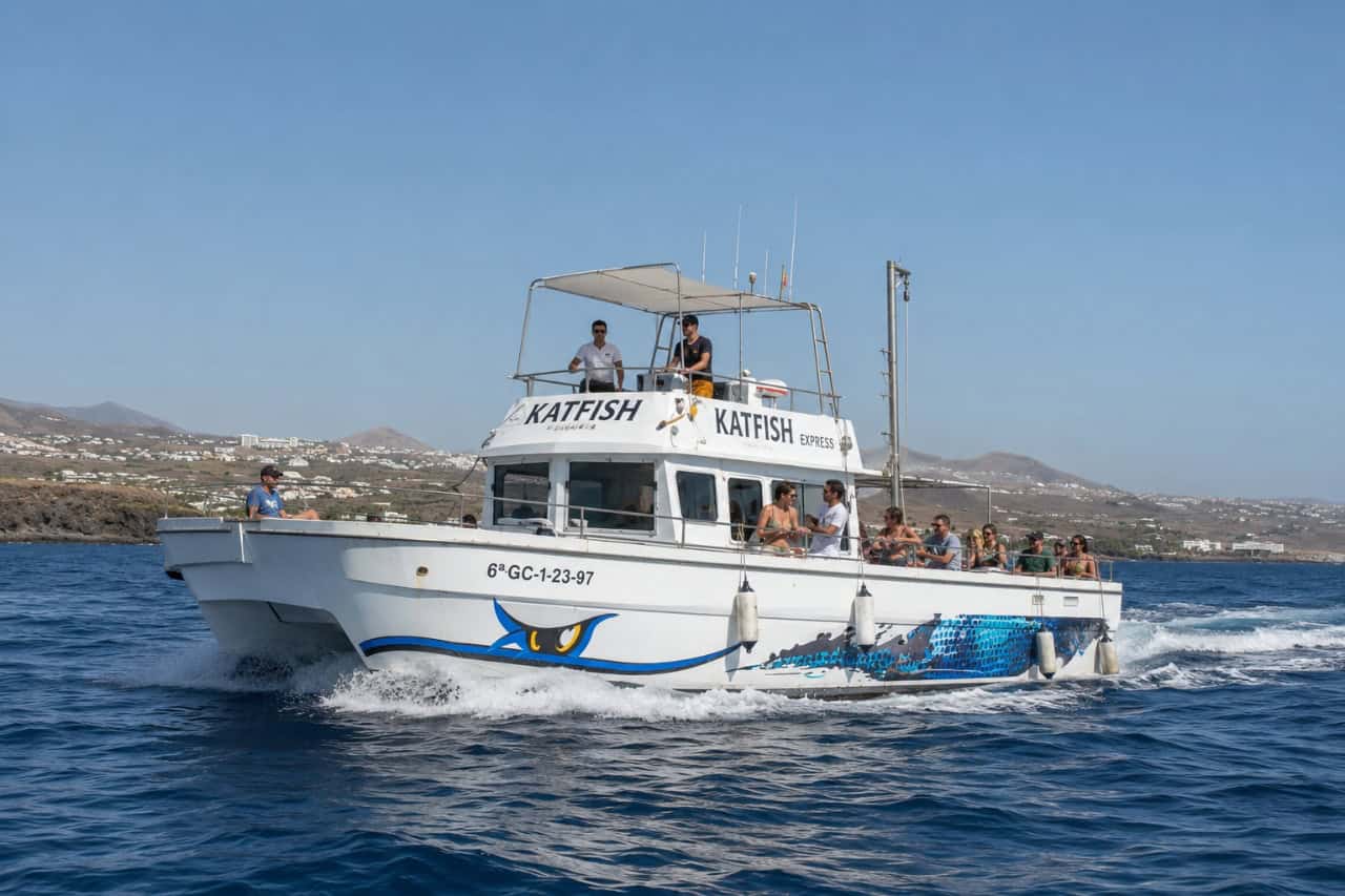 Katfish Express excursion boat cruising along the coast of Lanzarote with passengers on board, offering fishing trips and sightseeing against a backdrop of volcanic landscapes and the Atlantic Ocean.