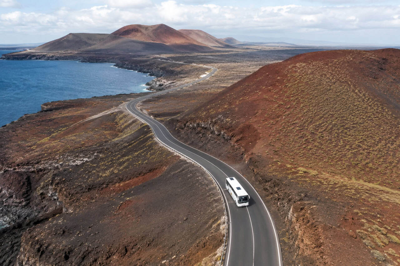 Drone view of a tour bus driving along Lanzarote's volcanic coastline with turquoise Atlantic waters and dramatic island landscapes.