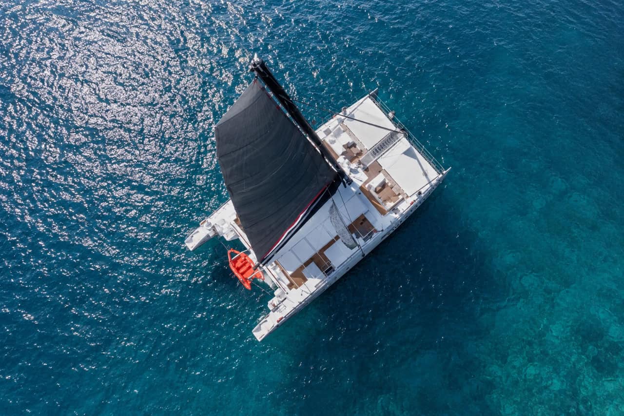 Aerial view of a luxury white catamaran with a black sail cruising over the crystal-clear turquoise waters of Tenerife, offering an exclusive private sailing experience.