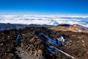 Hiker walking along the rocky trail to the summit of Mount Teide above the clouds, with volcanic landscapes and the Teide crater visible during a guided hike with cable car access.