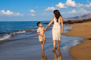 Mother and son walking hand in hand along a peaceful sandy beach in Tenerife during a golden-hour family photoshoot, with the ocean and volcanic coastline in the background.