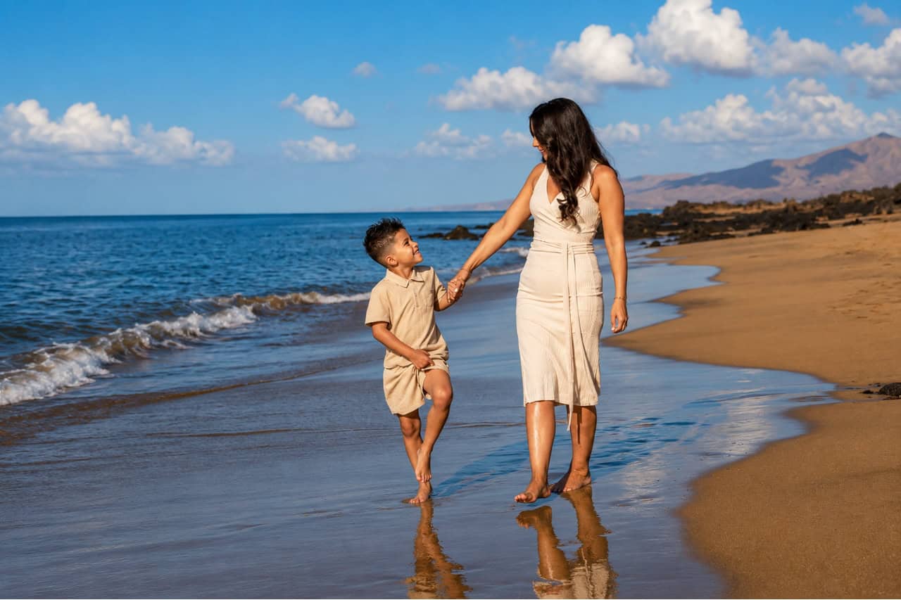 Mother and son walking hand in hand along a peaceful sandy beach in Tenerife during a golden-hour family photoshoot, with the ocean and volcanic coastline in the background.