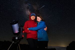 Couple posing beside a telescope during a stargazing experience under a clear, star-filled night sky with a visible shooting star overhead.