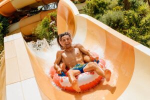 Two visitors riding an inflatable tube down a high-speed water slide at Siam Park in Tenerife, splashing through a sharp curve surrounded by tropical gardens.