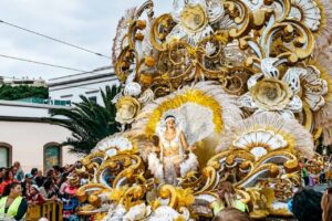 Carnival queen wearing an elaborate white and gold costume on a decorated parade float during the Tenerife Carnival, surrounded by spectators and festive celebrations.
