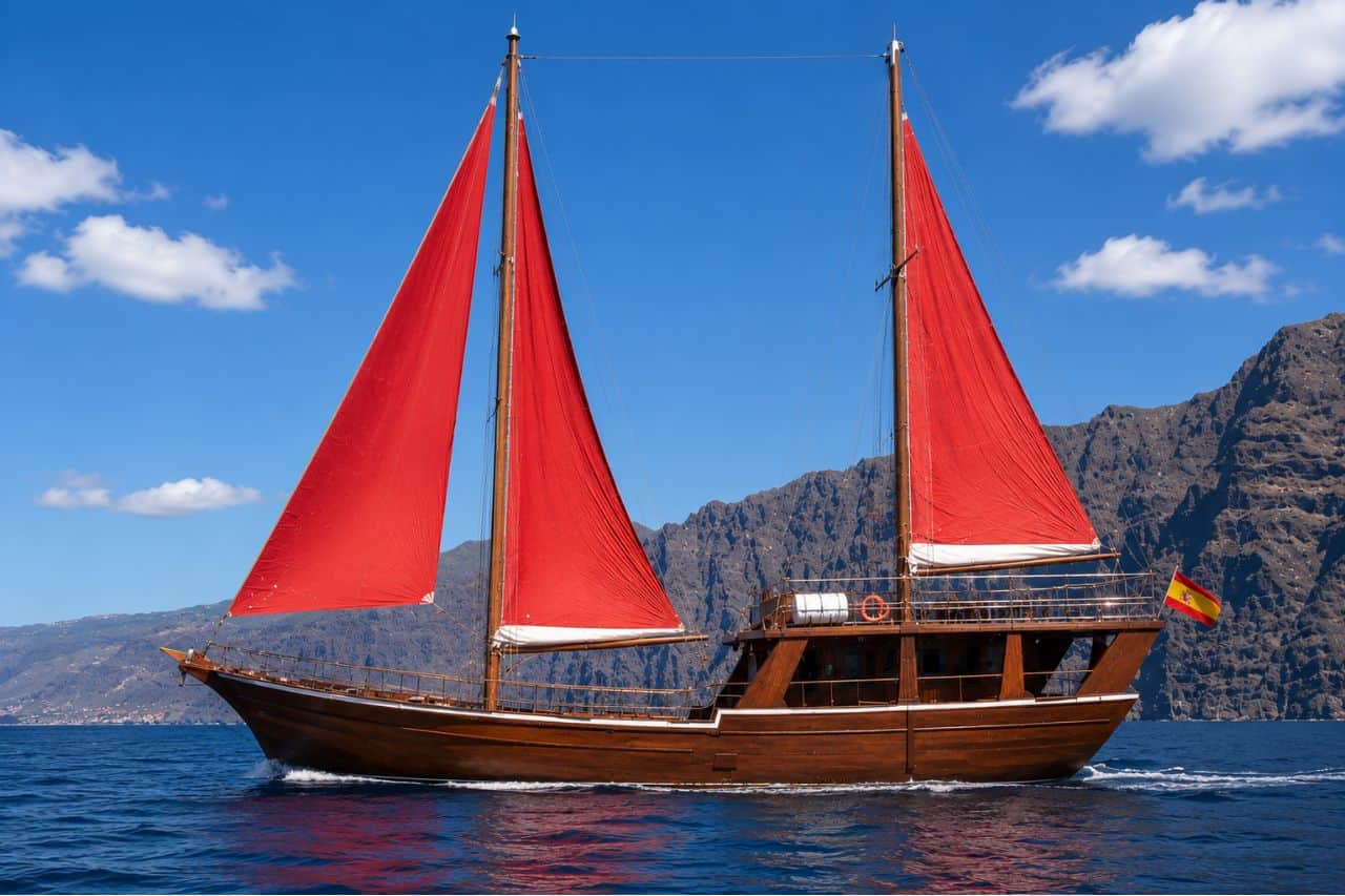 Traditional oriental-style sailing ship anchored off the cliffs of Tenerife during a whale watching cruise, with guests swimming in the Atlantic Ocean and enjoying lunch on board.