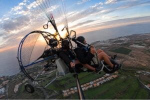 Two people enjoying a tandem paragliding flight over Tenerife at sunset, with panoramic views of the coastline, countryside and mountains below.