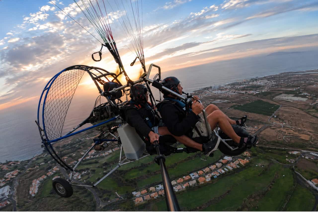 Two people enjoying a tandem paragliding flight over Tenerife at sunset, with panoramic views of the coastline, countryside and mountains below.