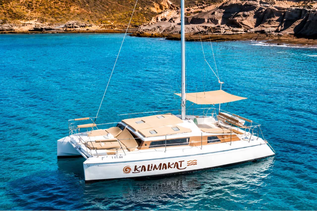 White 11-metre catamaran anchored in crystal-clear turquoise waters off the Tenerife coastline with dramatic volcanic cliffs in the background.
