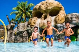 Tres niños corriendo por una piscina poco profunda frente a la gigantesca cascada en forma de calavera y la zona infantil de temática pirata en Aqualand Maspalomas, en Gran Canaria