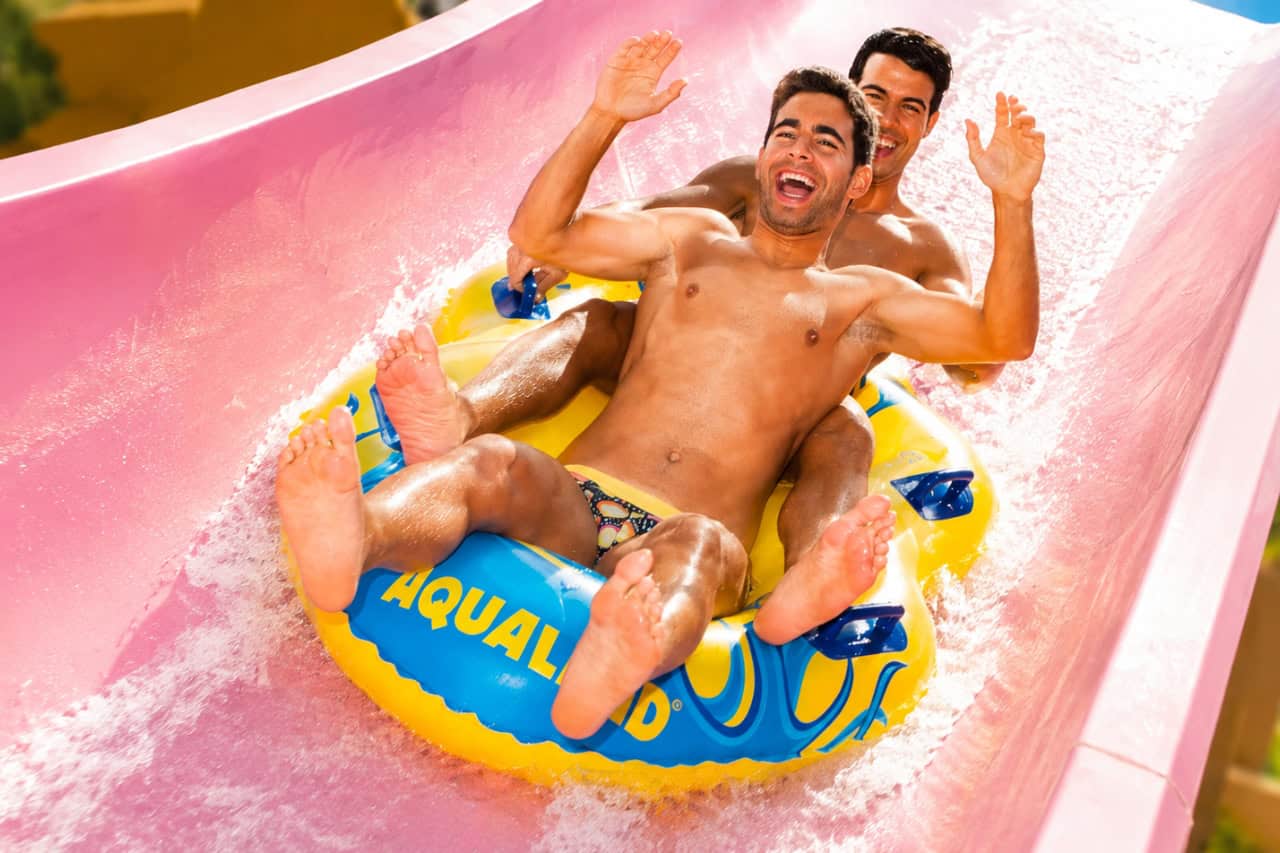 Two friends enjoying a fast inflatable raft ride down a pink water slide at Aqualand Maspalomas, surrounded by splashing water on a sunny day