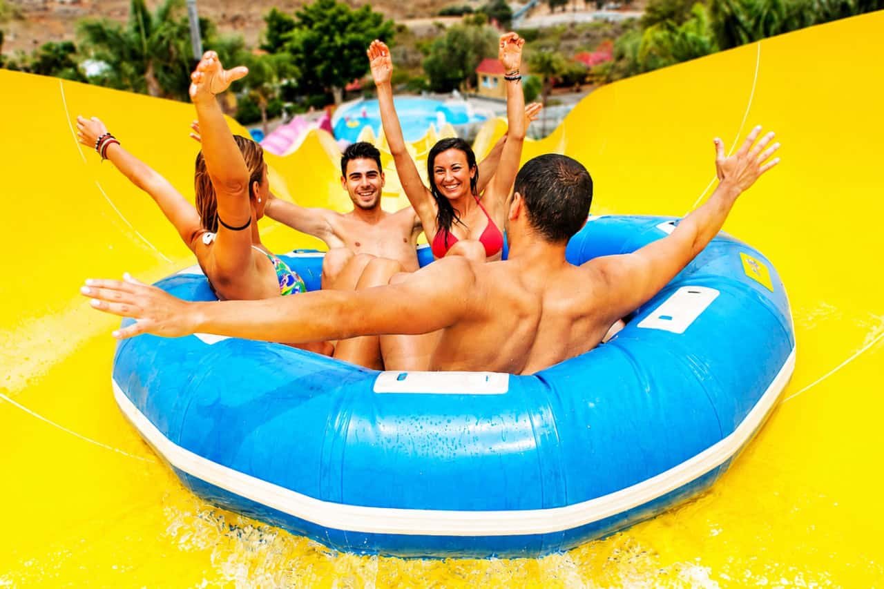 Group of four friends riding together in a large blue inflatable raft down a bright yellow water slide at Aqualand Maspalomas in Gran Canaria
