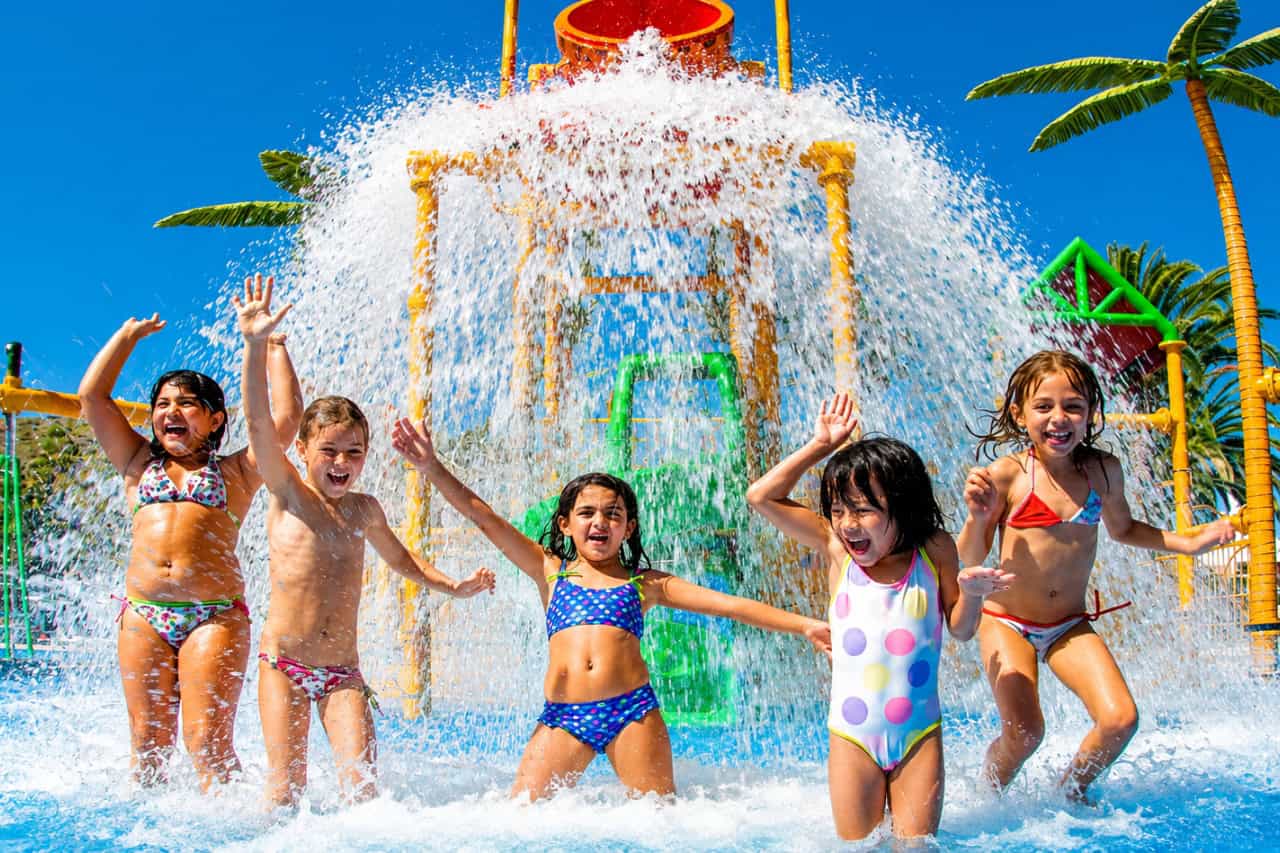 5 children playing and laughing under a large tipping water bucket, surrounded by colourful splash attractions and palm trees at Aqualand Maspalomas waterpark
