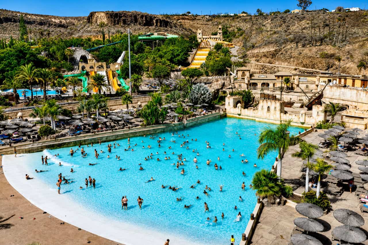 Panoramic view of the large wave pool at Aqualand Maspalomas, with swimmers, palm trees, sunbathing areas and water slides surrounded by Gran Canaria’s rocky landscape