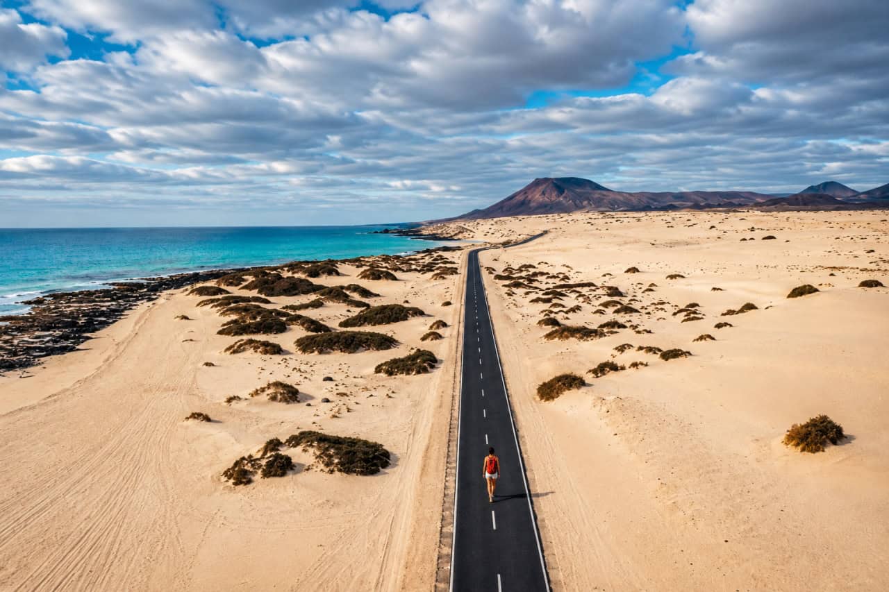 Aerial view of the road through Corralejo Natural Park with golden sand dunes, turquoise coastline, and volcanic mountains in Fuerteventura.