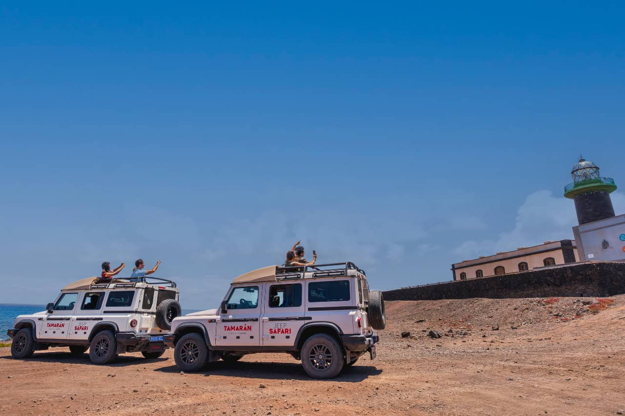 Two Jeep Safari vehicles with travellers exploring the rugged coastline near a lighthouse in Fuerteventura.