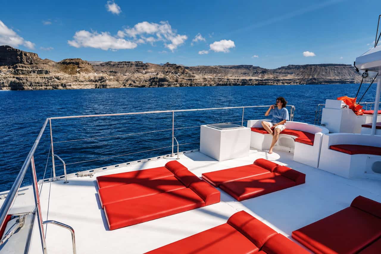 Huésped relajándose en la amplia cubierta de un catamarán privado de lujo durante un alquiler de catamarán privado en Gran Canaria, con dramáticos acantilados costeros de fondo.