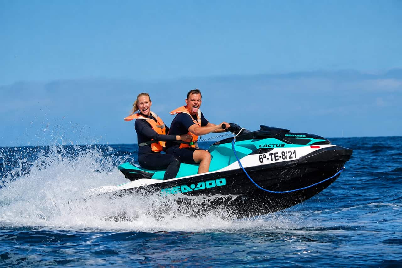 Couple riding a turquoise Sea-Doo during a guided jet ski safari in Tenerife, speeding across the Atlantic Ocean with sea spray behind them.
