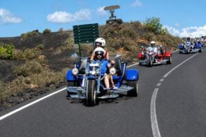 Skupina jazdcov jazdiaca na modrých a červených trojkolkách (trikes) sopkovou krajinou Národného parku Timanfaya počas organizovanej výhliadkovej jazdy na trojkolkách po Lanzarote.