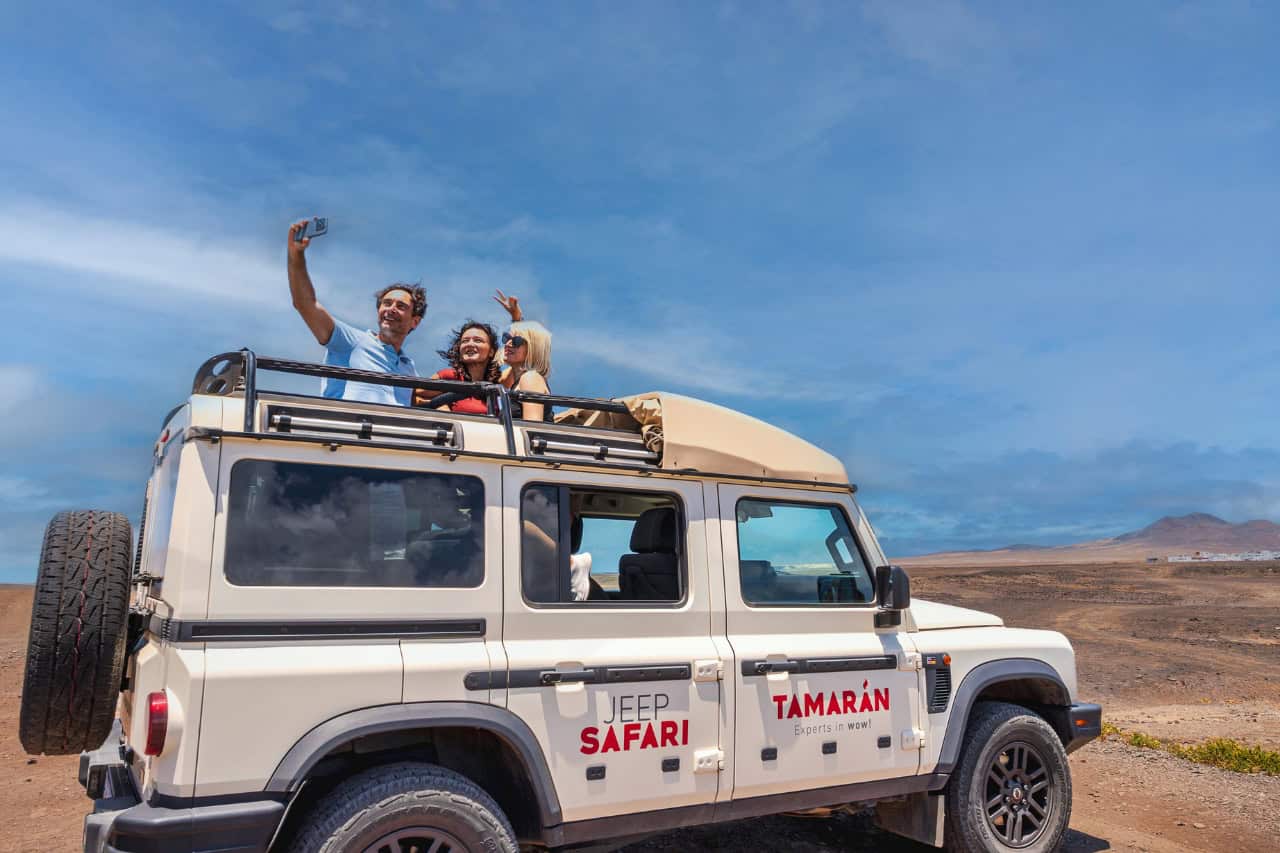 Three people taking a selfie from an open-top 4x4 Jeep while exploring the off-road volcanic landscapes of Fuerteventura.