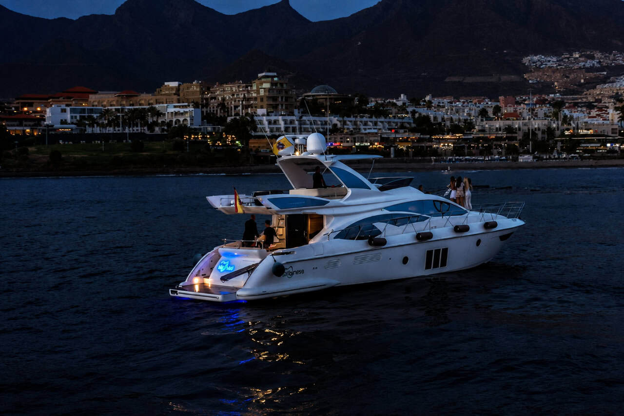 Luxury white Tigresa yacht cruising off Costa Adeje at dusk, with Tenerife’s coastline, resort lights and dark mountain backdrop visible behind the boat.