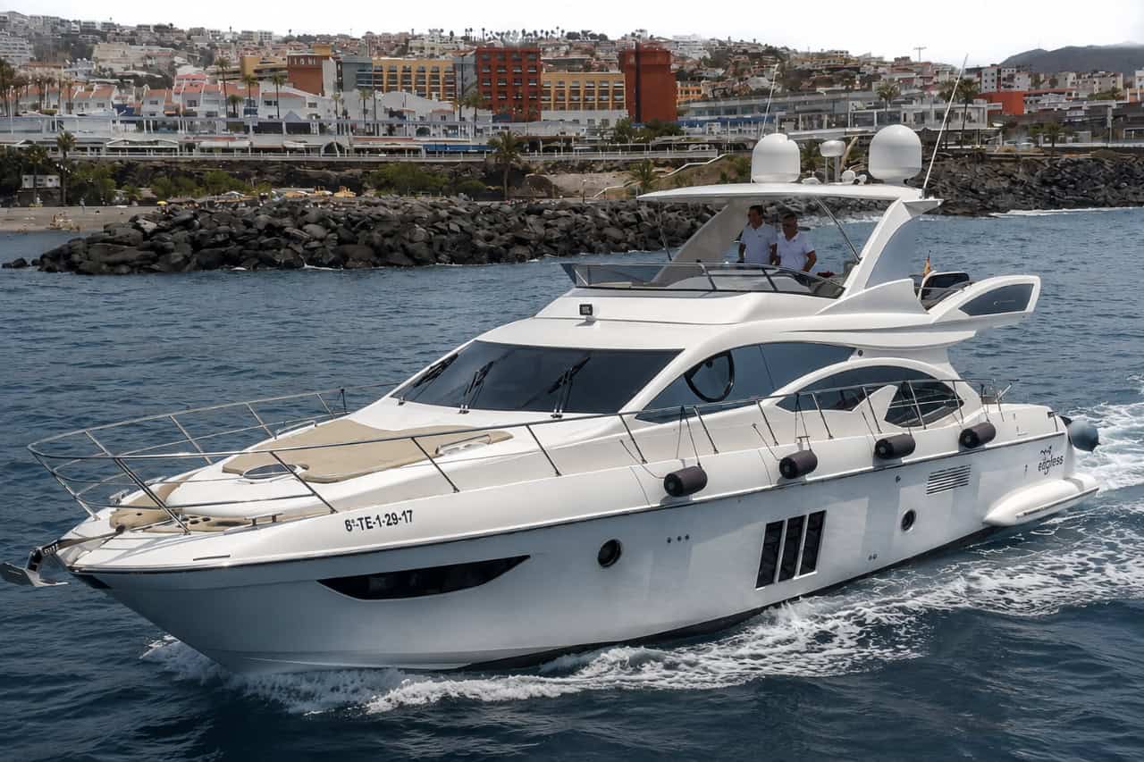 White Tigresa luxury yacht cruising through blue water near Costa Adeje, with two crew members on the flybridge and resorts behind it.