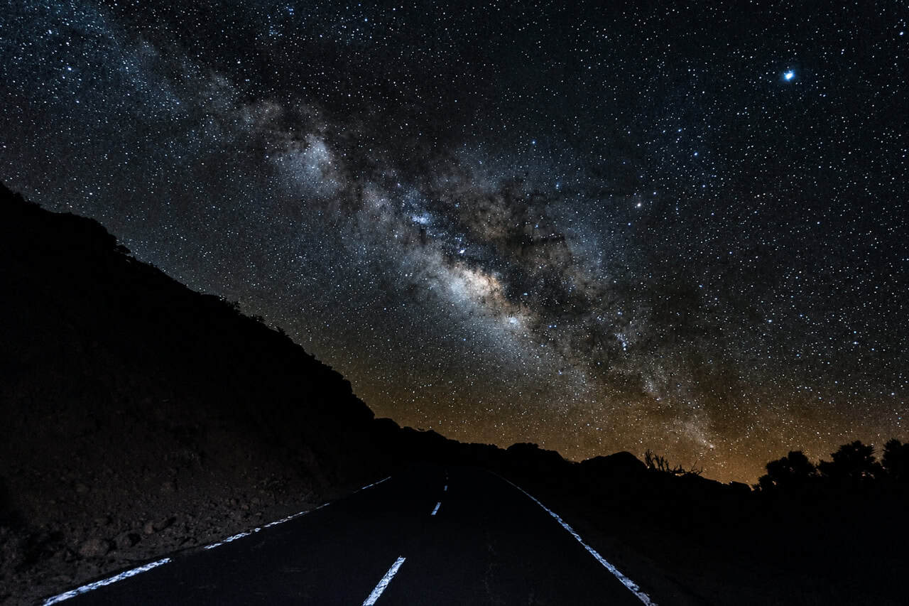 Milchstraße, die während einer Sternennacht über einer abgelegenen Bergstraße auf Gran Canaria leuchtet, umgeben von dunklem Vulkangelände.