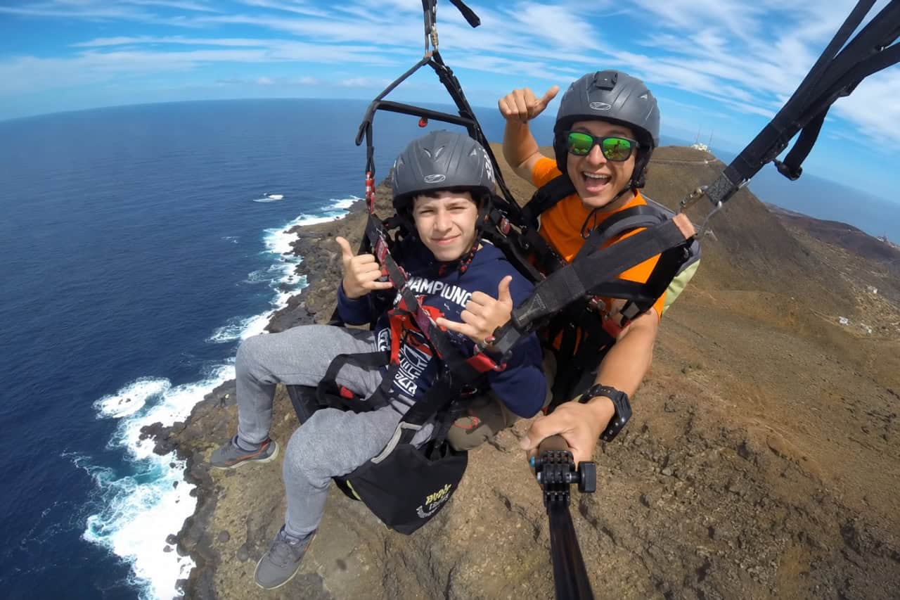Passagier und Tandem-Gleitschirminlehrer lächeln und zeigen den Daumen nach oben, während sie über der dramatischen Küste von Gran Canaria fliegen. Beide tragen Helme und vollständige Sicherheitsgurte, wobei während des Gleitschirmflugs die Vulkanlandschaft, Klippen und der Atlantische Ozean unter ihnen zu sehen sind.