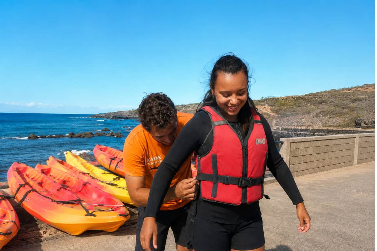 Kayaking instructor helping a guest correctly secure her red life jacket before a kayak tour in Tenerife, with colourful kayaks and the rocky coastline visible behind them. The image highlights the personal safety assistance and preparation provided before entering the water.