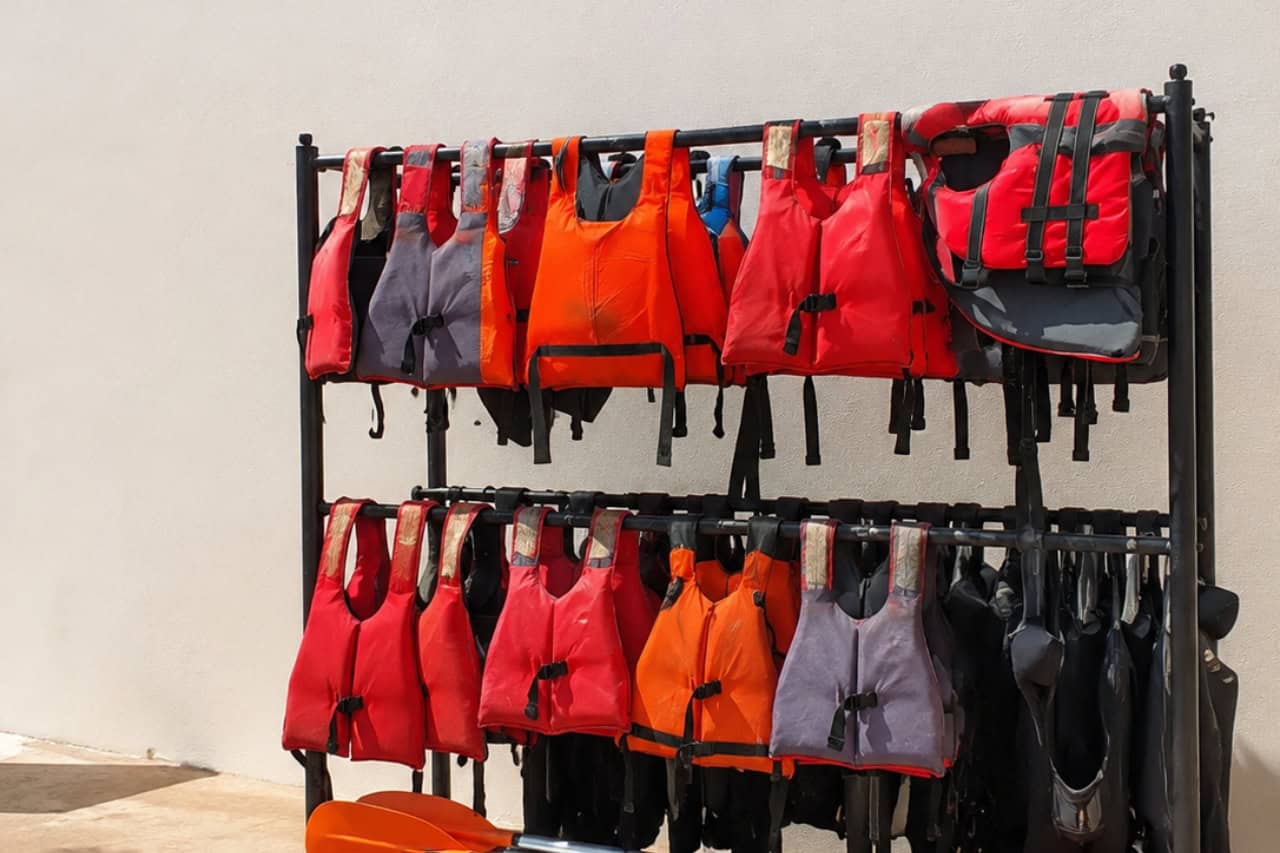 Selection of red, orange and grey life jackets arranged on an equipment rack with kayak paddles ready for guests before a kayaking tour in Tenerife. The image shows the safety equipment provided for participants before heading onto the water.