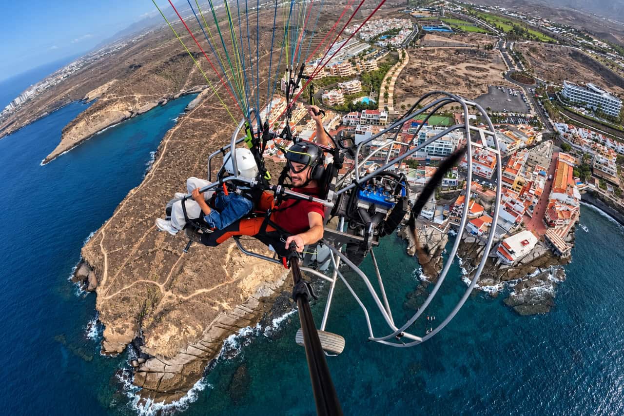 Tandem paratrike flying above Tenerife’s rocky coastline with panoramic views of the Atlantic Ocean and coastal resorts.