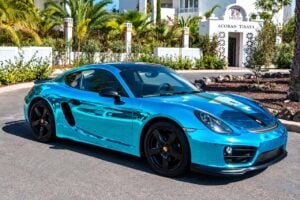 Blue Porsche 718 Cayman with black alloy wheels and racing stripe parked in a tropical setting, available for luxury car rental in Tenerife.