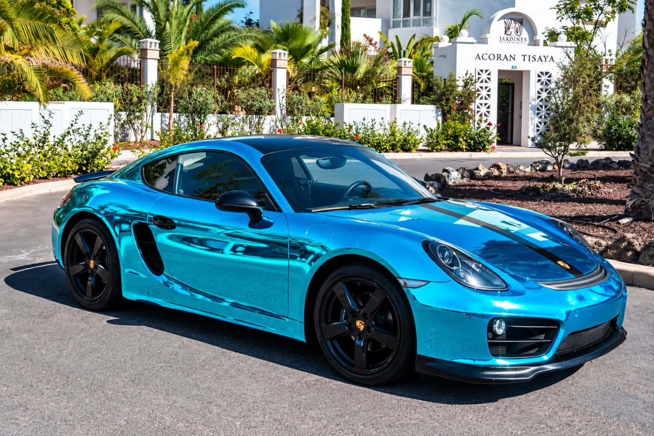 Blue Porsche 718 Cayman with black alloy wheels and racing stripe parked in a tropical setting, available for luxury car rental in Tenerife.