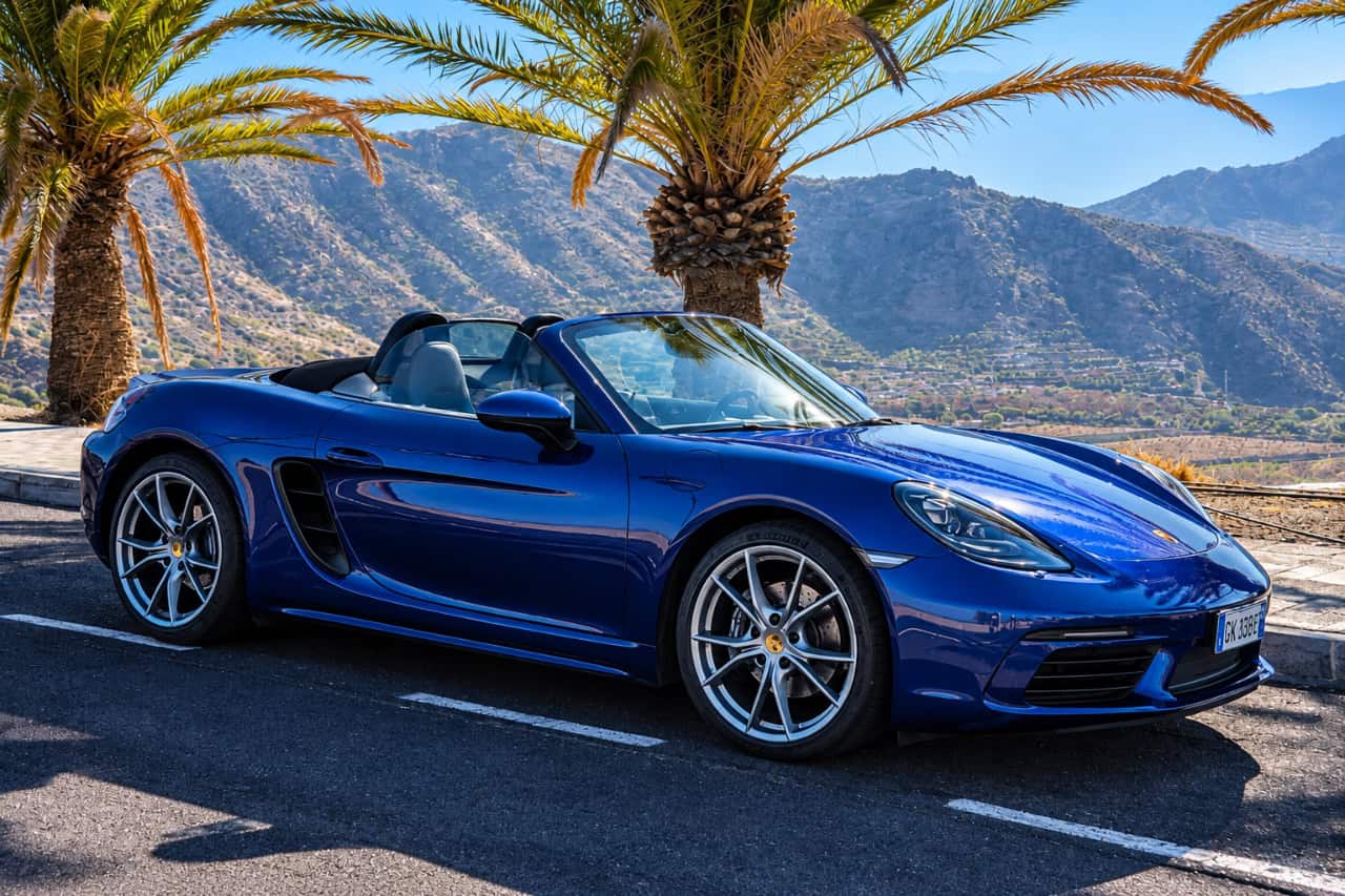 Blue Porsche 718 Boxster convertible with the roof down, parked beneath palm trees with Tenerife’s mountain landscape in the background.