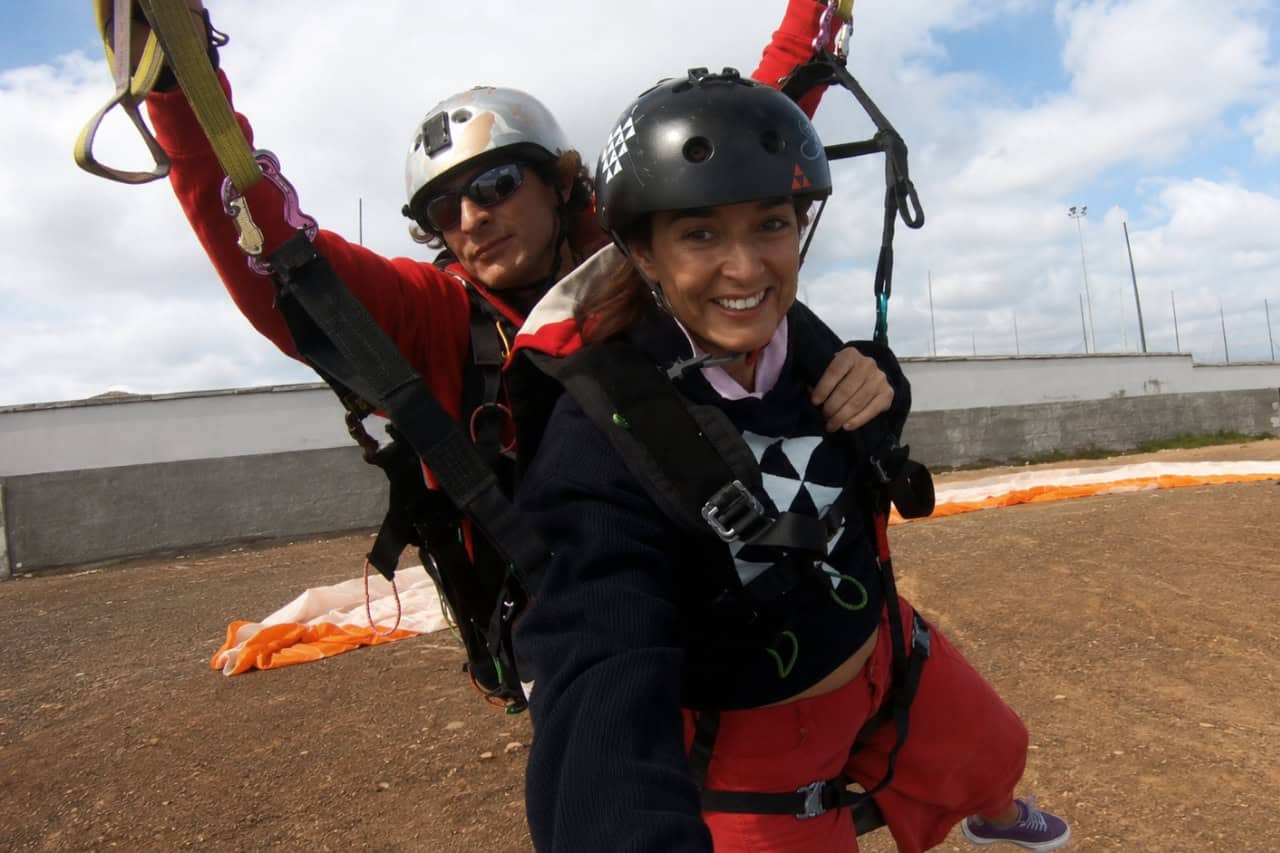 Lächelnde Kundin, die sich auf Gran Canaria auf den Start zu einem Tandem-Gleitschirmflug vorbereitet, während sie in einem Vollsitzgurt und mit Helm an ihrem Fluglehrer gesichert ist. Der Gleitschirmkappe liegt hinter ihnen am Startplatz ausgebreitet, während sich die beiden auf den Beginn des Fluges vorbereiten.