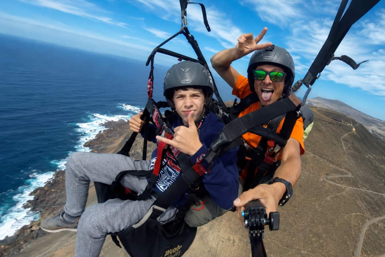 Jinquerer Passagier genießt einen Tandem-Gleitschirmflug auf Gran Canaria mit einem erfahrenen Fluglehrer, beide gesichert in Helmen und Gurtzeugen hoch über der zerklüfteten Vulkanküste und dem Atlantischen Ozean. Das Paar posiert während des Fluges für die Kamera und zeigt die unterhaltsame und abenteuerliche Seite eines Gleitschirm-Erlebnisses auf Gran Canaria.