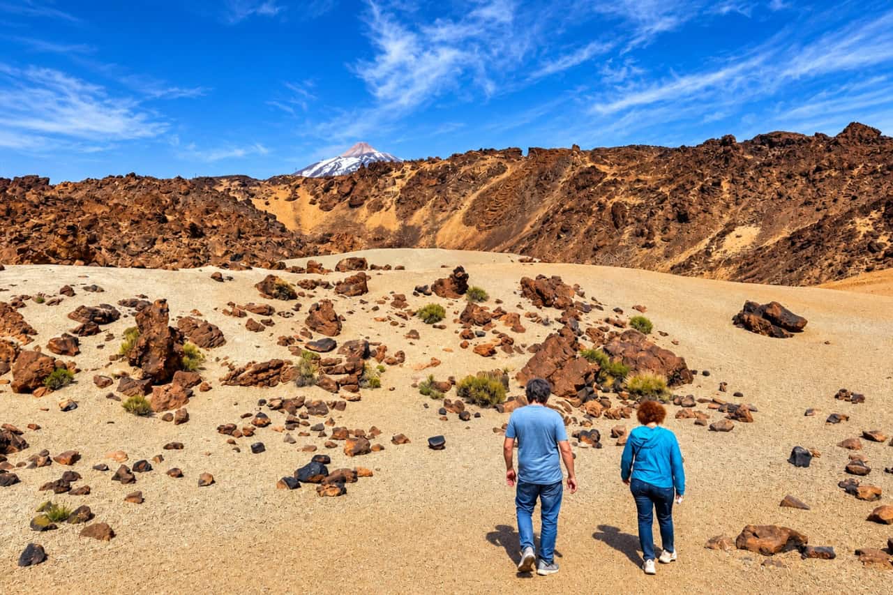 Two visitors walking across pale volcanic sand and dark lava rock in Teide National Park