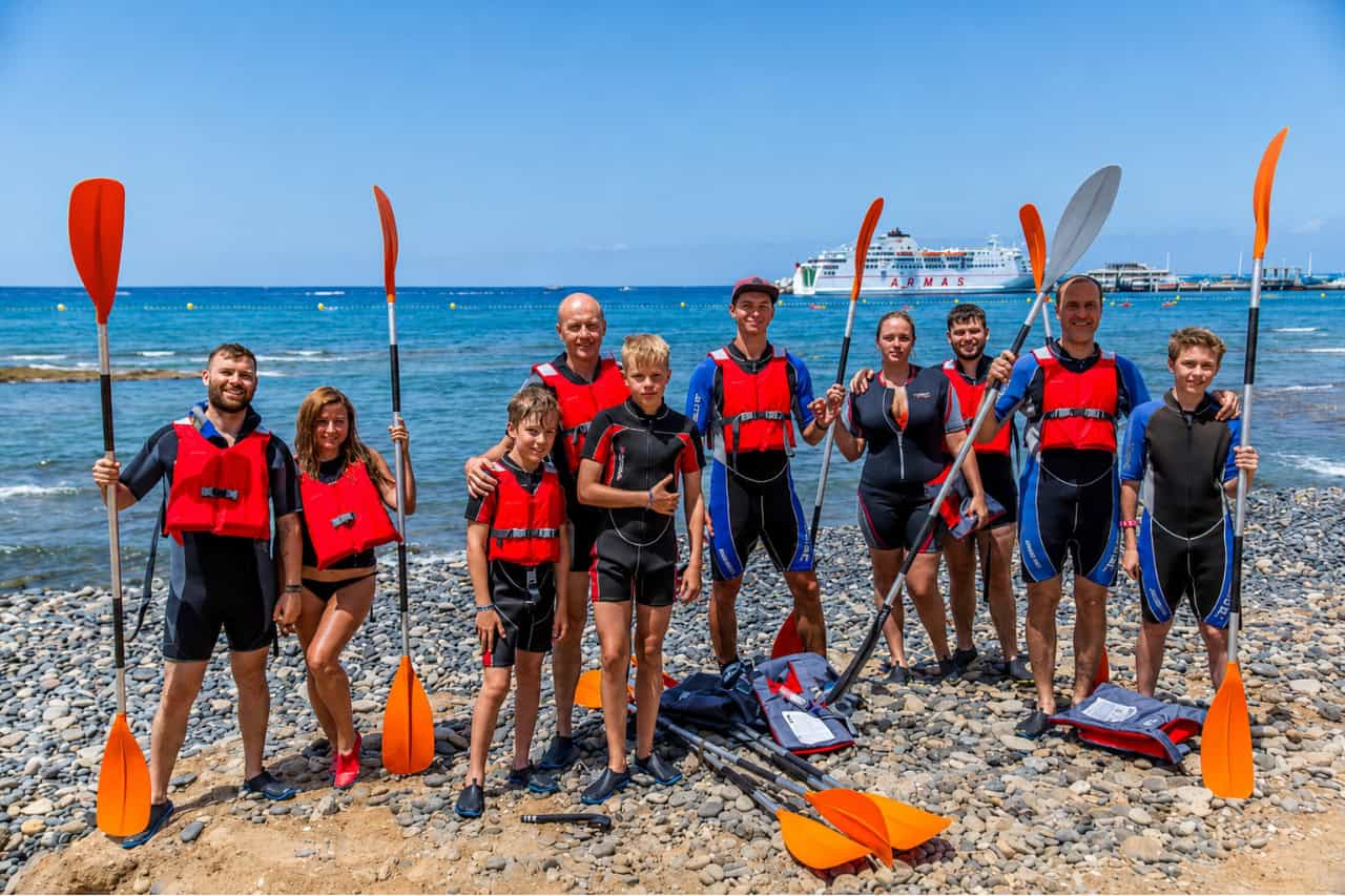 Group of adults and children wearing life jackets and holding kayak paddles together on a rocky Tenerife beach before or after a guided kayaking excursion, showing the family-friendly and small-group atmosphere of the experience.