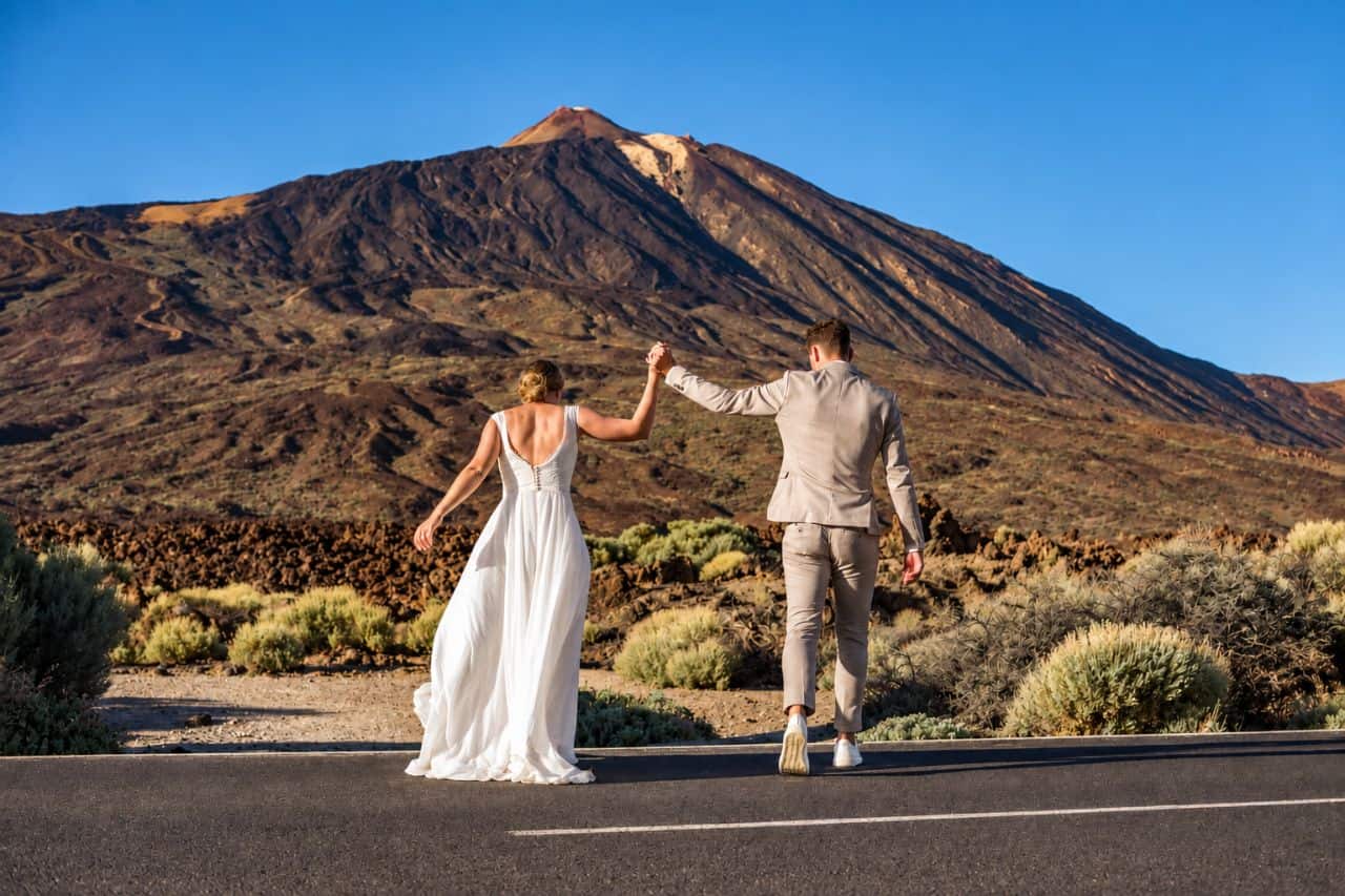 Een koppel in trouwjurken loopt met elkaar de hand in hand, met de Mount Teide achter zich, Tenerife