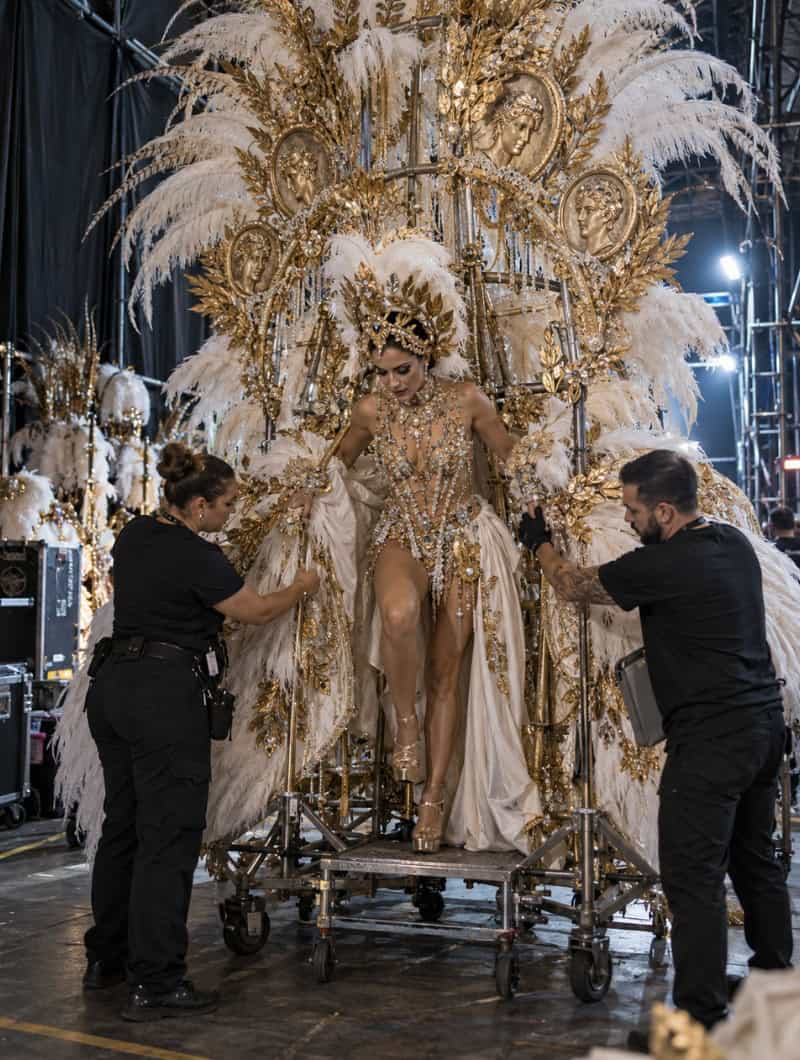 Impressie van een Tenerife Carnavalkoningin die backstage in haar gigantische witte en gouden verenjurk stapte op een wieltrapport, met twee medewerkers die hielpen
