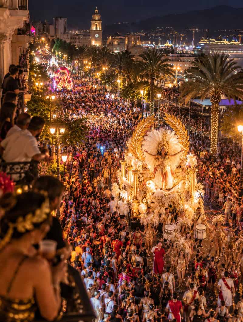 Impresie van het carnaval van Santa Cruz de Tenerife 's nachts, gezien vanaf een balkon, een gouden drijvende vloot en tienduizenden mensen in kostuums die de straat naar de haven vullen