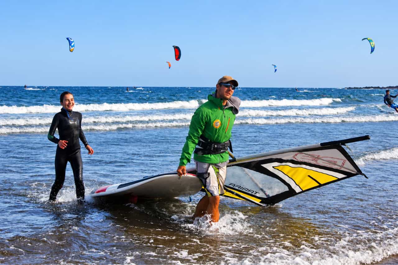 Windsurf instructor carrying a windsurf board and sail from the water at El Médano beach in Tenerife, with another participant and kitesurfers in the background.